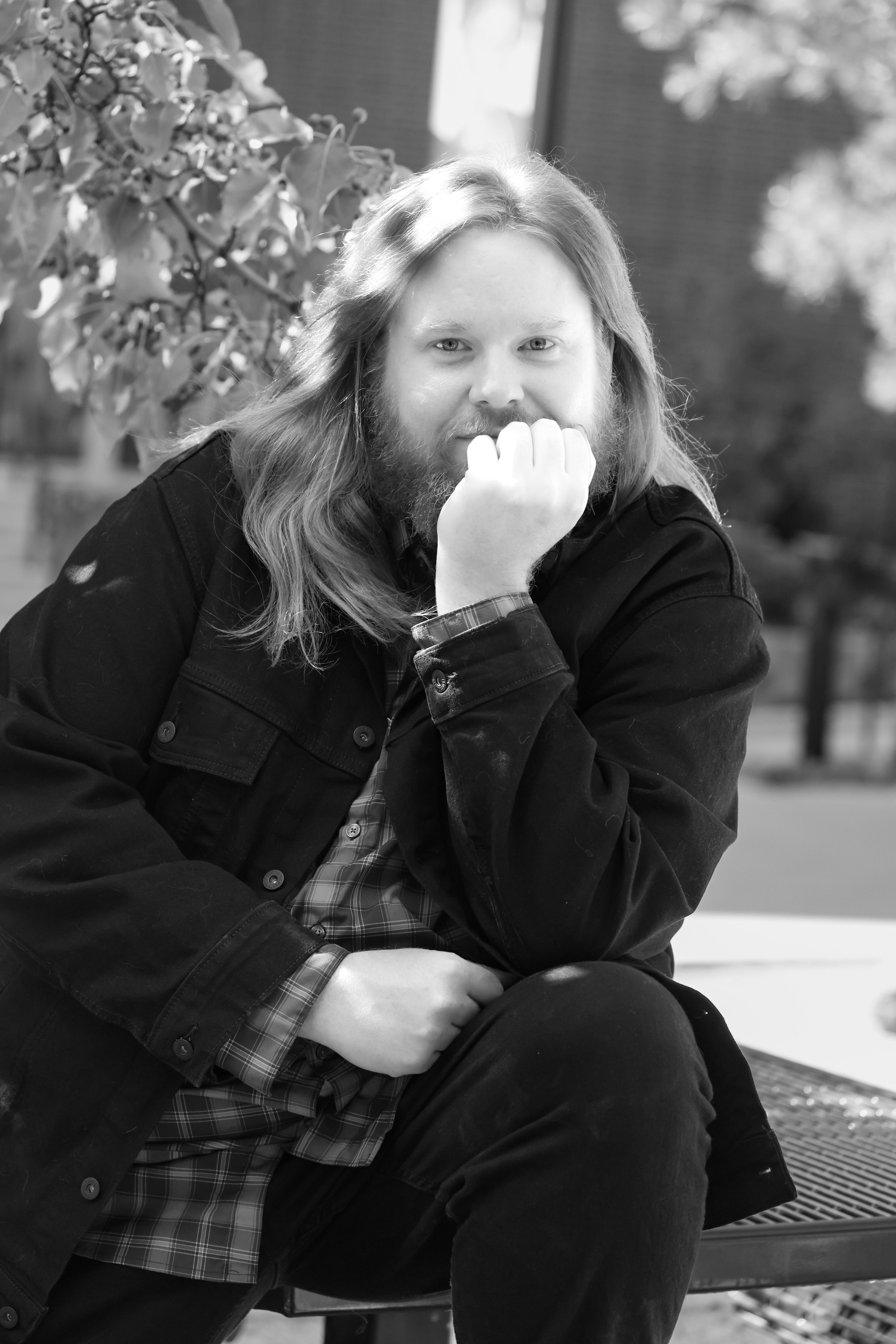 Christopher J Baker in black and white, sitting in a park in a denim jacket, resting his chin on his palm.